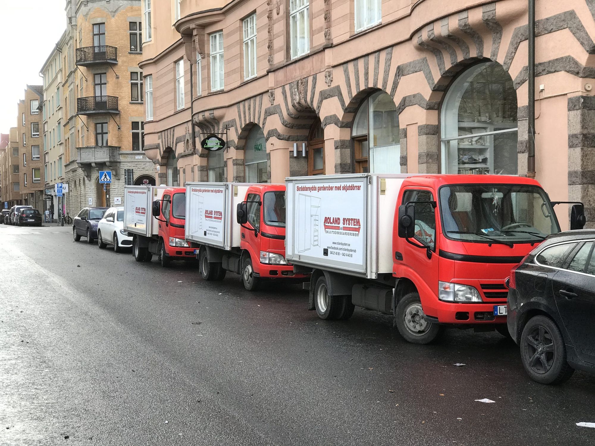 Tre röda Roland System-lastbilar parkerade på rad på en stadsgata framför ett äldre flerbostadshus.