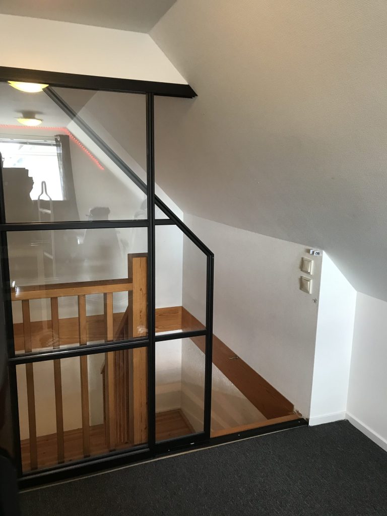 Glasvägg med svart ram under ett snedtak bredvid ett trapphus med träräcke.