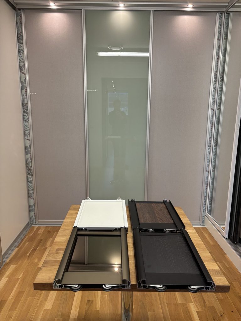 Cross-sections of sliding door frames with rollers on a table in the showroom, in front of grey fabric doors and a frosted glass door.
