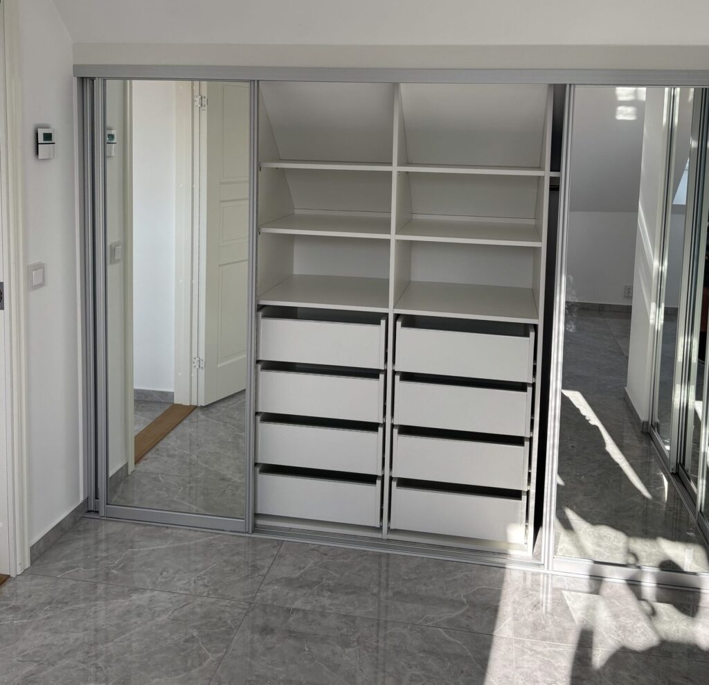 White shelves and four drawers fitted under a sloped ceiling, with mirror sliding doors on the sides.